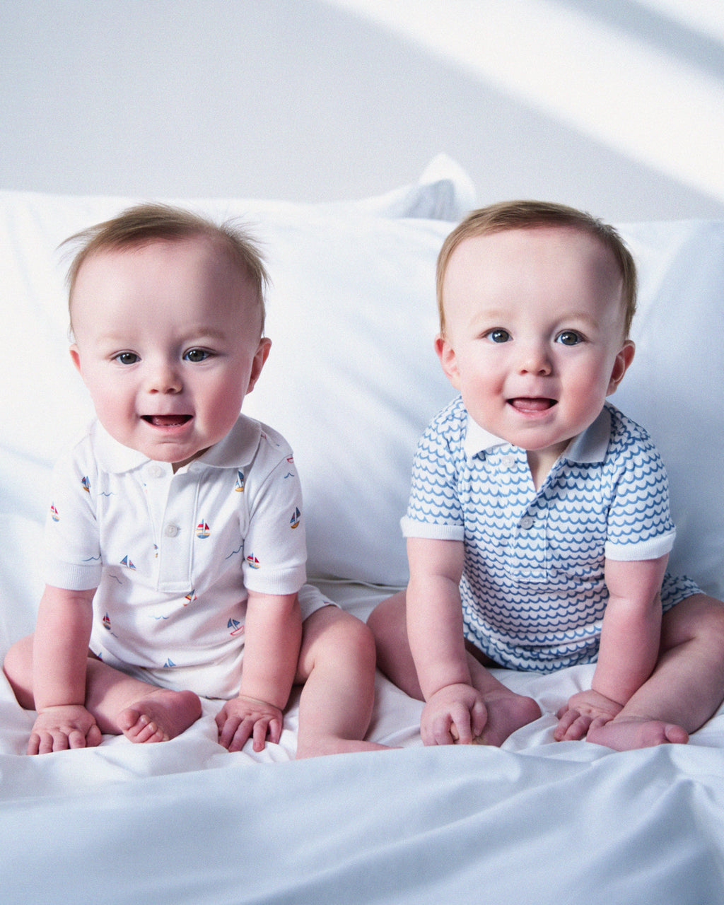 Two babies smile on a bed; one wears a white Pima cotton shirt with sailboat prints, while the other dons Petite Plumes Babys Pima Louis Play Romper in La Mer. The background features white bedding evoking the tranquility of the sea.