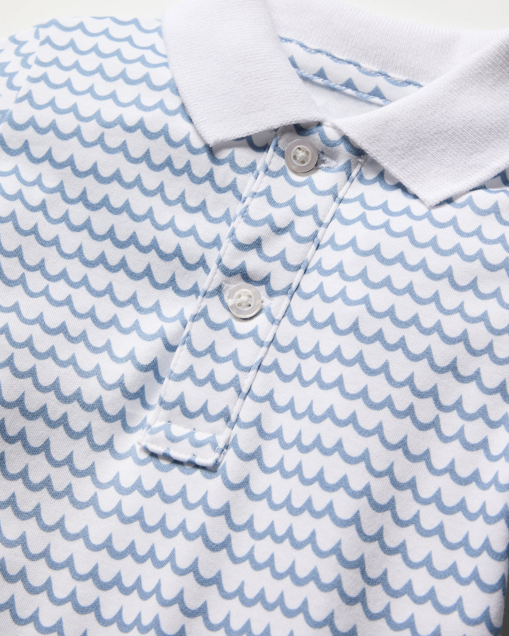 Close-up of Petite Plumes Babys Pima Louis Play Romper in La Mer, made from soft Pima cotton. It showcases a vibrant blue wave pattern on white, detailed with a crisp white collar and a buttoned placket with three matching buttons.