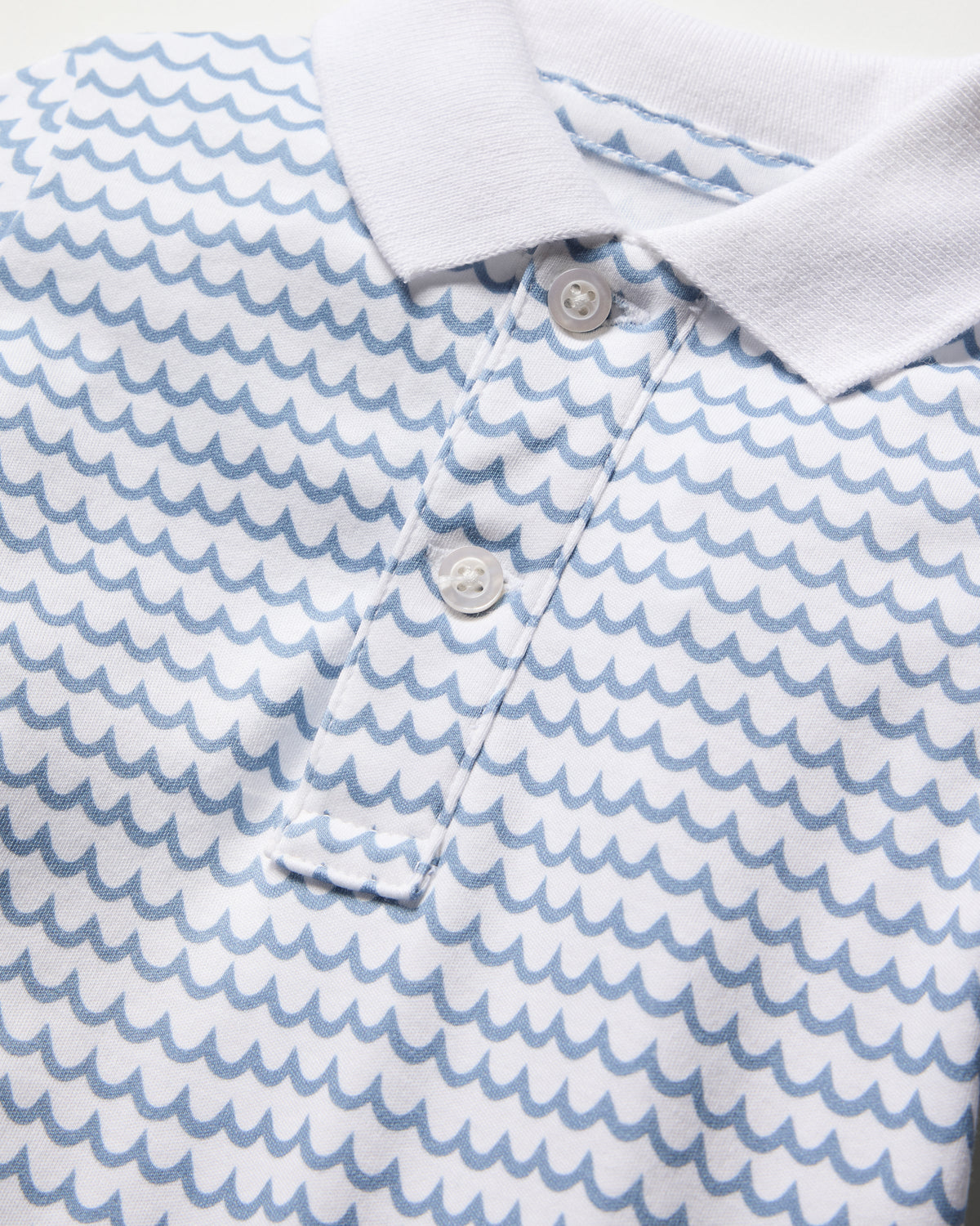 Close-up of Petite Plumes Babys Pima Louis Play Romper in La Mer, made from soft Pima cotton. It showcases a vibrant blue wave pattern on white, detailed with a crisp white collar and a buttoned placket with three matching buttons.