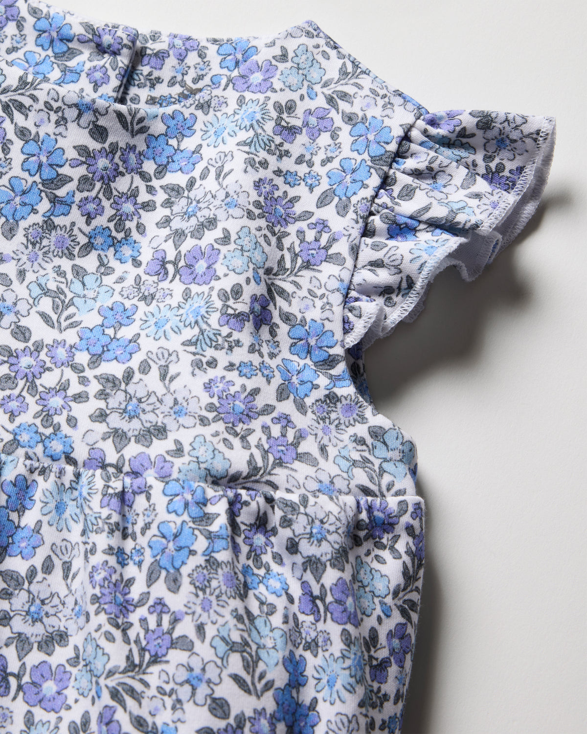 Close-up of Petite Plumes Baby Girl Pima Play Romper in Fleur dAzur, featuring azure blue and purple flowers on white. The fabric showcases a gathered design with ruffled sleeves, highlighting its vintage print charm.