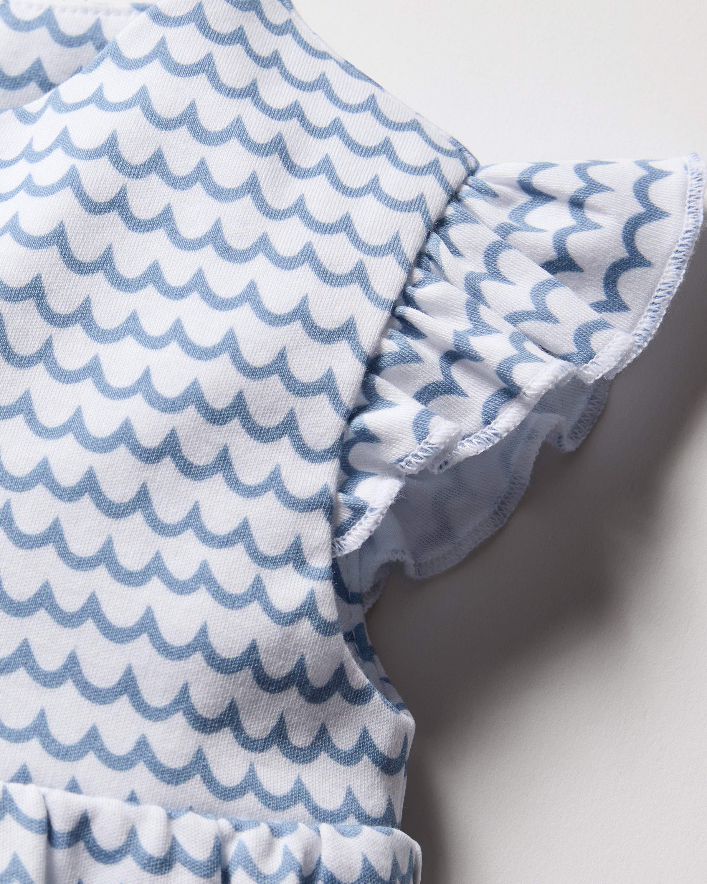 Close-up of Petite Plumes Babys Pima Isla Play Romper in La Mer, showcasing a blue wave pattern on white pure Pima cotton. Features include short frilly sleeves and neat stitching.
