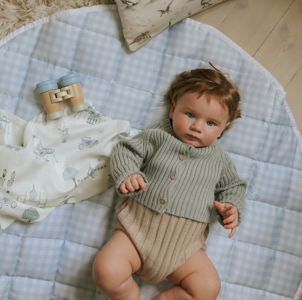 Baby lying on a round quilted light blue gingham play mat with a wooden toy and patterned blanket on a light wood floor