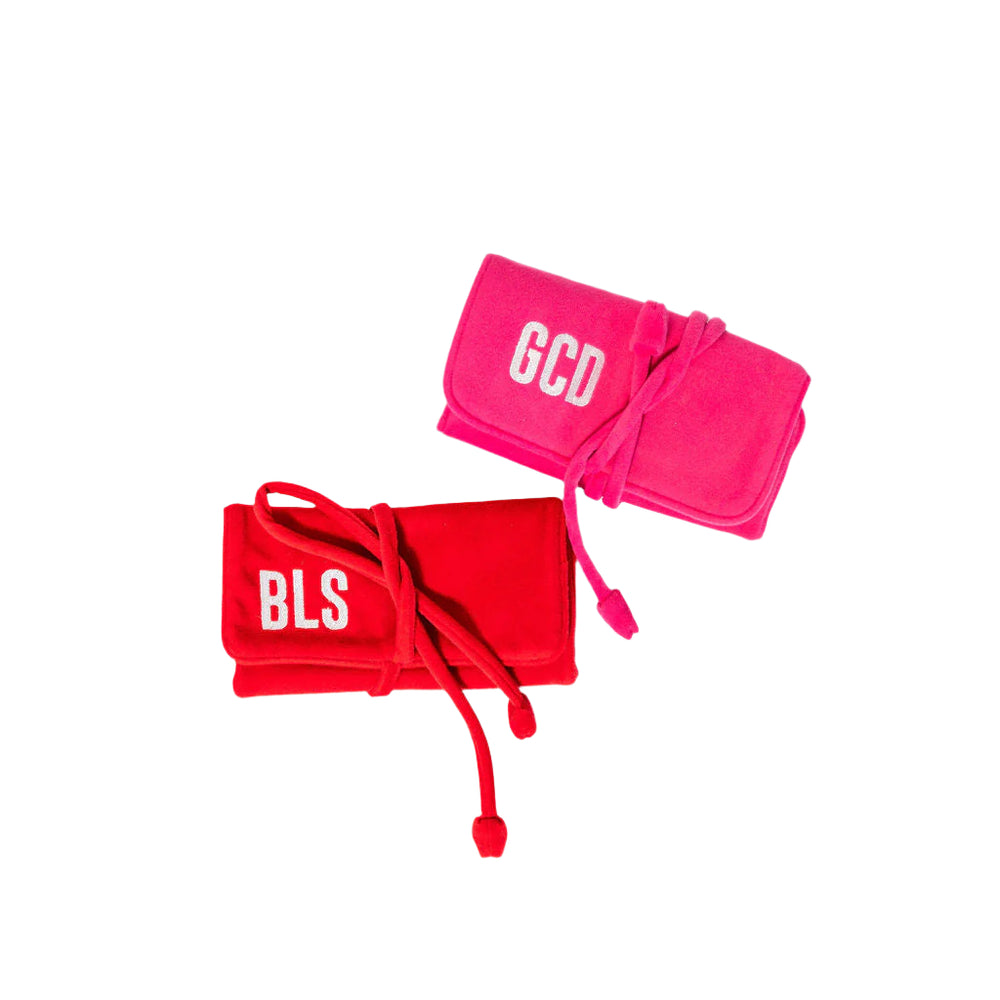 Angled view of two fabric jewelry rolls (red and hot pink) showcasing tie closures and embroidered monograms ‘BLS’ and ‘GCD’ on a white background