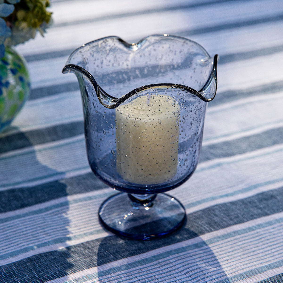 Front view of Provence 5.5 inch Hurricane in Chambray with ruffled rim and pedestal base, holding a candle on plaid fabric