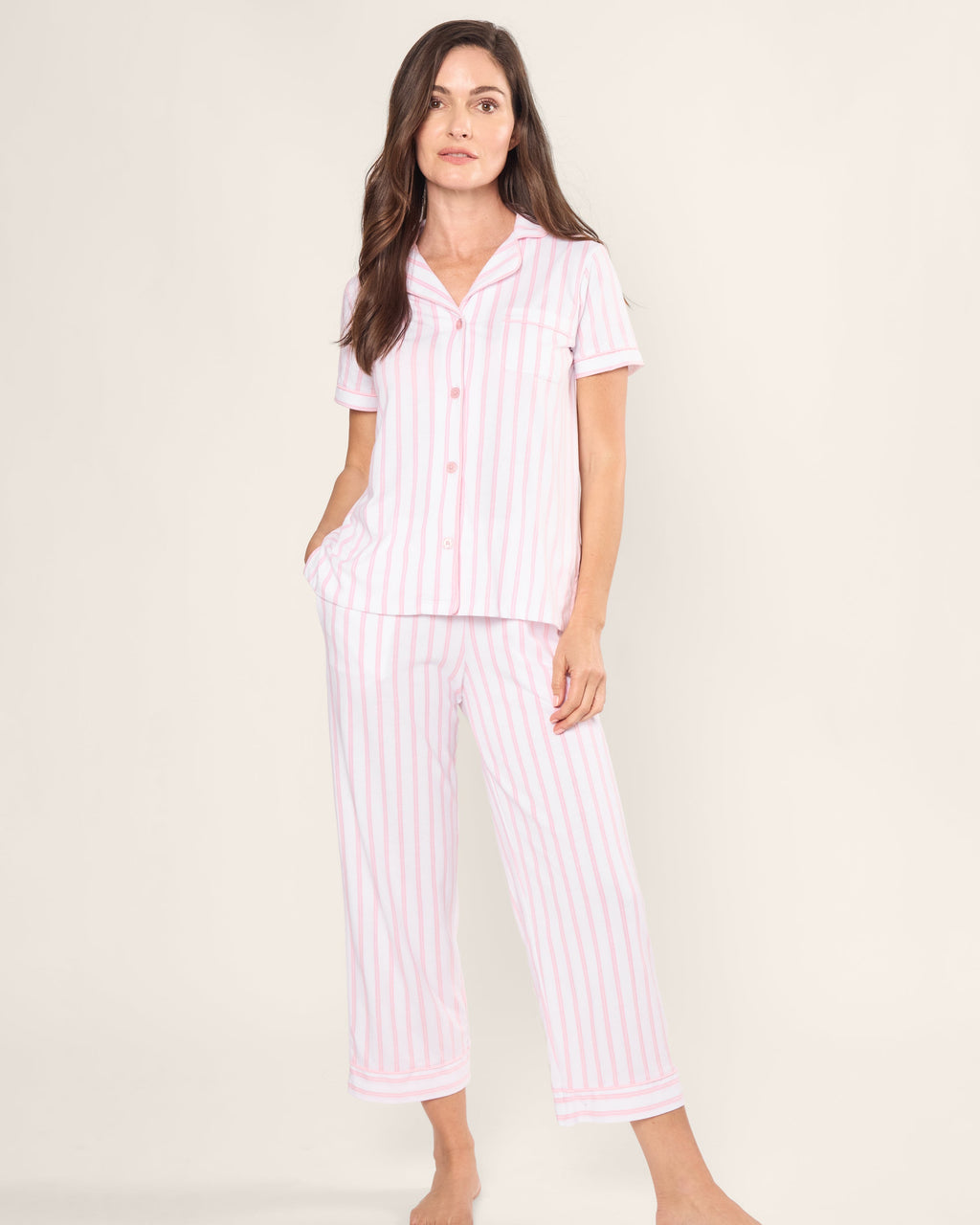 A woman stands barefoot with one hand in a pocket, wearing Petite Plumes Womens Pima Crop Set in Pink and White Stripe. The soft Pima cotton set includes a button-up shirt with short sleeves and matching pants, all accentuated by the plain background for a timeless elegance.