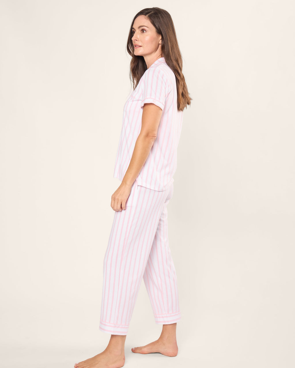 A woman stands barefoot in profile wearing Petite Plumes Womens Pima Crop Set in a pink and white stripe, featuring a luxurious vintage-style short-sleeved button-up shirt and cropped pants. She is set against a plain, light-colored background.