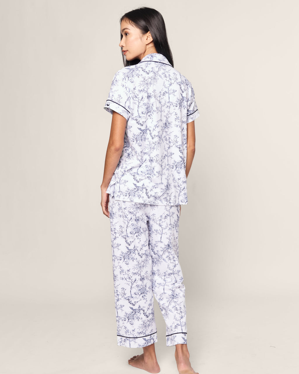 A woman with long dark hair faces away, modeling the Womens Pima Crop Set in Timeless Toile by Petite Plume. The light ensemble features a classic floral print with short sleeves and a collar top, paired with full-length pants against a plain, neutral background.