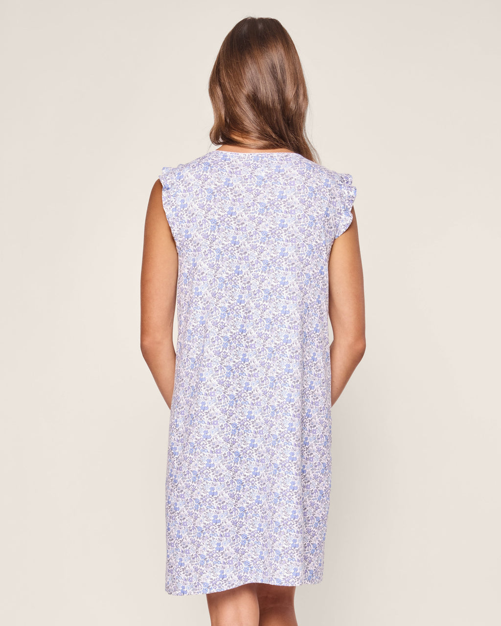 A person with long hair stands facing away, wearing a sleeveless Womens Pima Harper Nightgown in Fleur dAzur from Petite Plume, showcasing a delicate purple floral pattern on a light background that ends above the knees.
