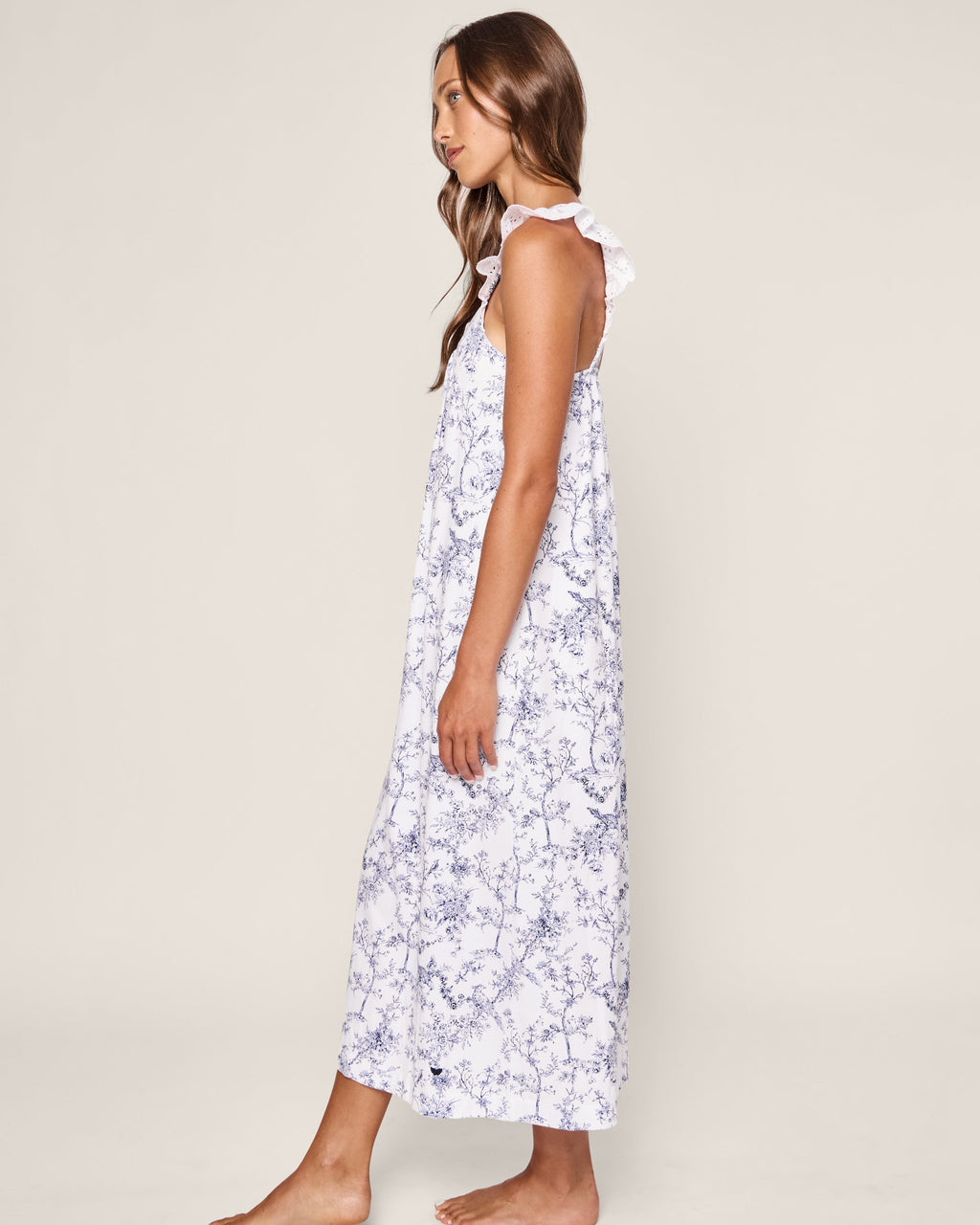 A woman with long hair stands barefoot against an off-white background, embracing her bedtime serenity. She wears Petite Plumes Womens Pima Clara Nightgown in Timeless Toile, a sleeveless ankle-length white dress with blue floral patterns and ruffled straps, facing sideways.