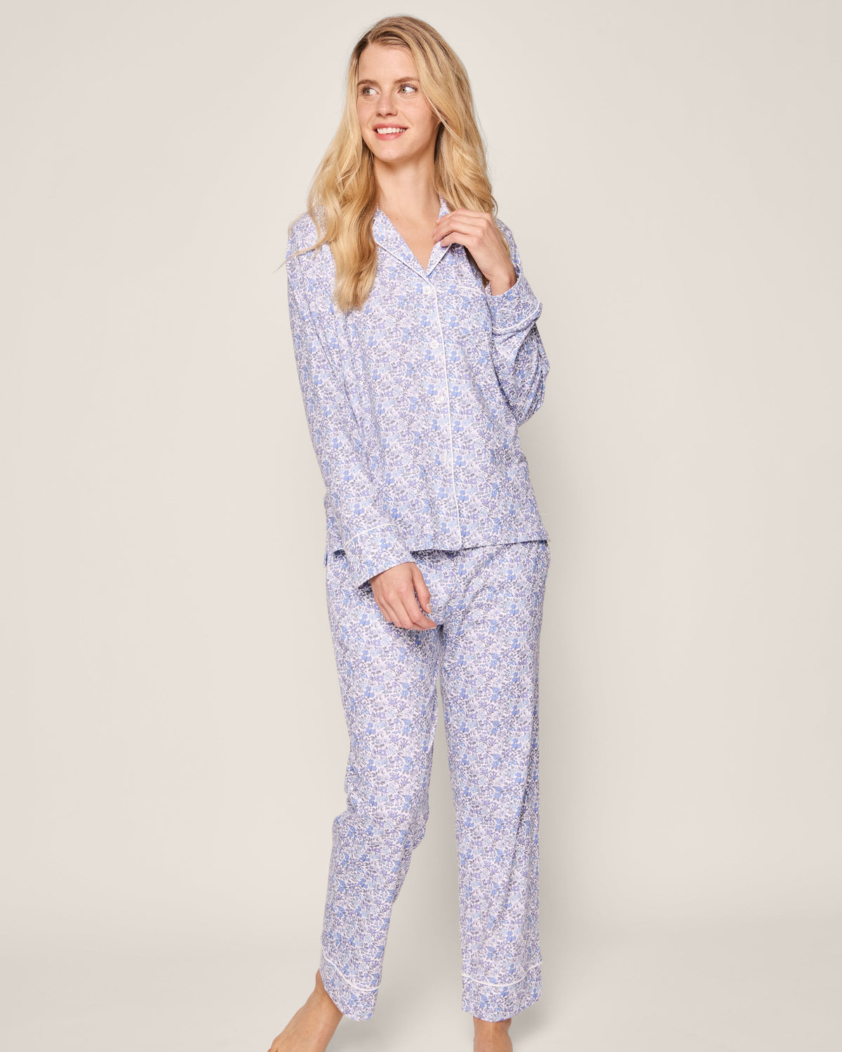 A person smiles while barefoot in Petite Plumes Womens Pima Pajama in Fleur dAzur, a vintage-inspired light purple floral set. Made from Peruvian Pima cotton, it features a long-sleeve button-up shirt with a collar and matching pants against a neutral background.