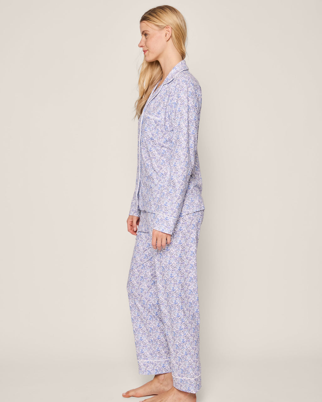 A person with long blonde hair stands barefoot in profile, wearing Petite Plumes Womens Pima Pajama in Fleur dAzur, featuring a vintage-inspired blue and white floral design. The background is plain and neutral.