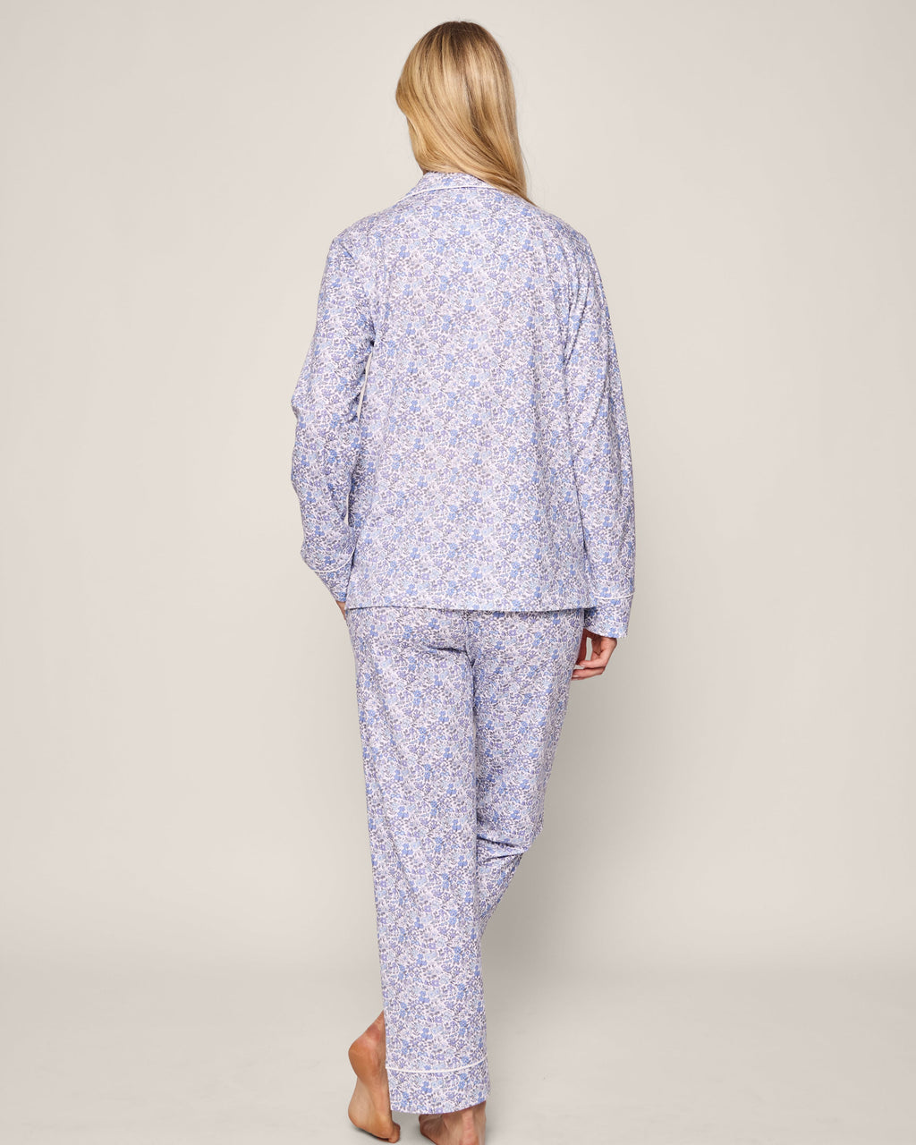 A person with long blonde hair stands barefoot, facing away, wearing the vintage-inspired Petite Plume Womens Pima Pajama in Fleur dAzur. The light blue patterned pajamas with long sleeves and pants create an elegant look against a plain background.
