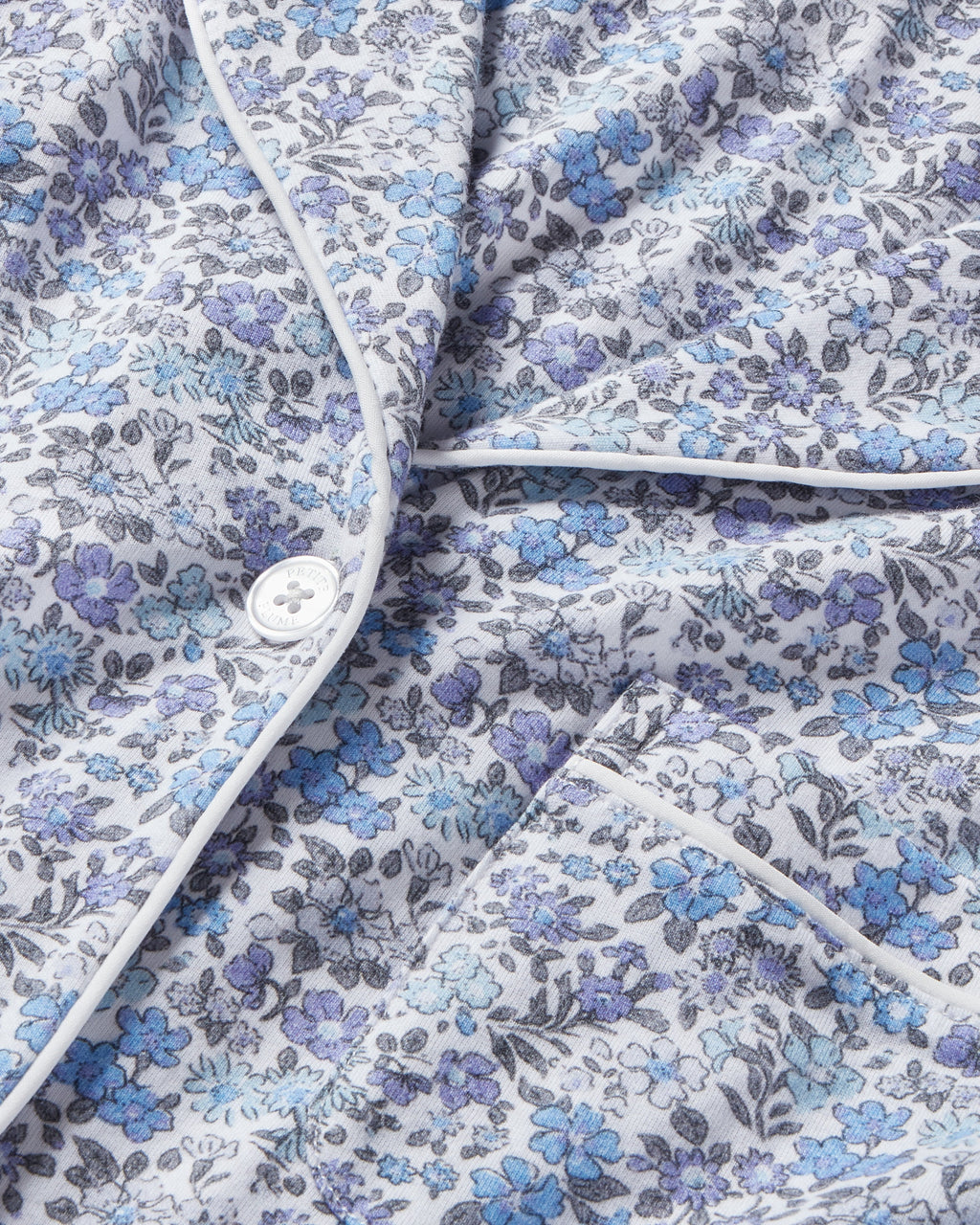 Close-up of Petite Plumes Womens Pima Pajama in Fleur dAzur, featuring a floral pattern in blue and purple on luxurious Peruvian Pima cotton. A white button, piping, and visible pocket enhance its vintage-inspired look against a soft grayish background.