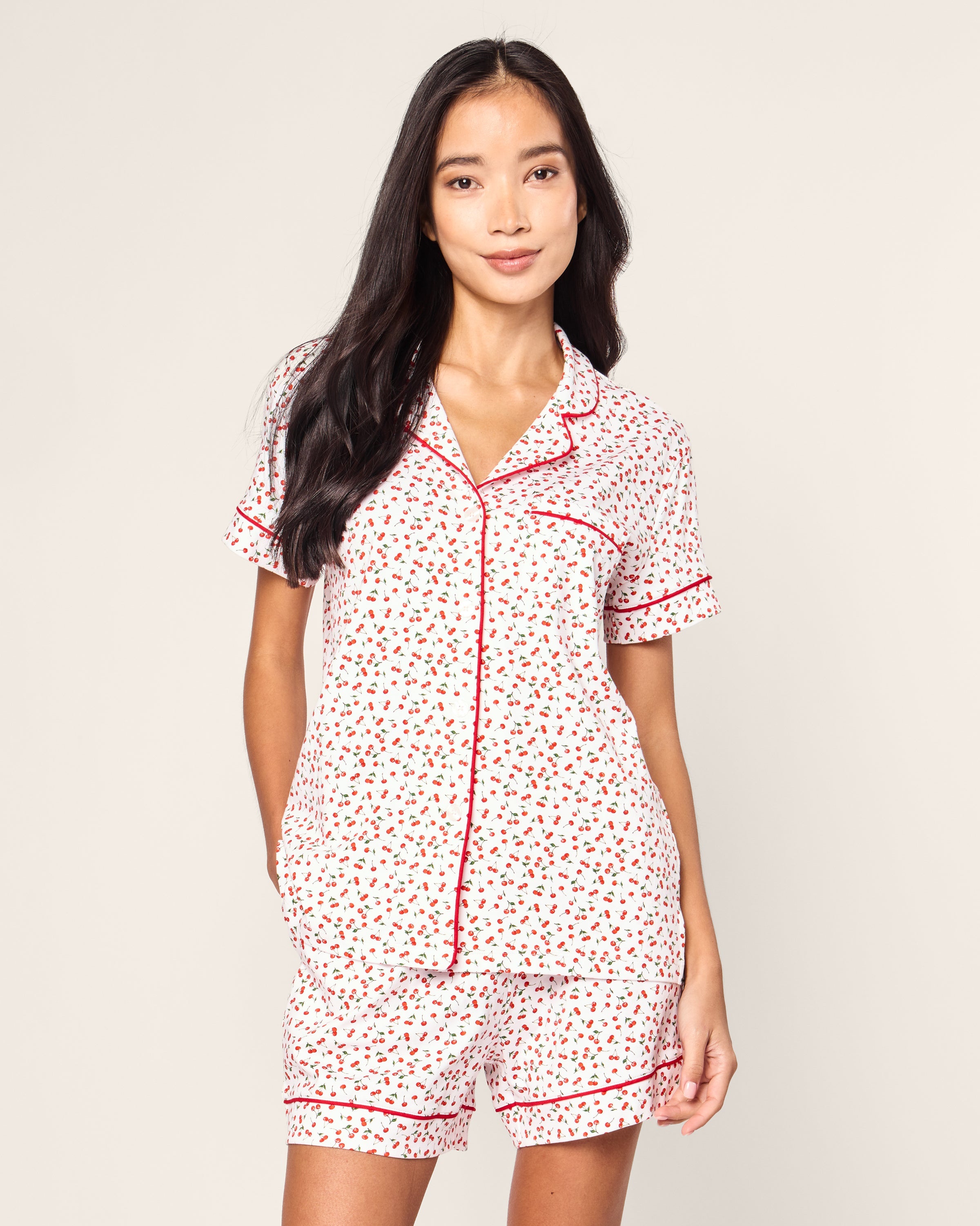 A person with long dark hair wears Petite Plumes Womens Pima Short Sleeve Short Set in Cherry Orchard, a white jersey knit pajama ensemble with a collared top, short sleeves, matching shorts, and red piping on a neutral background.