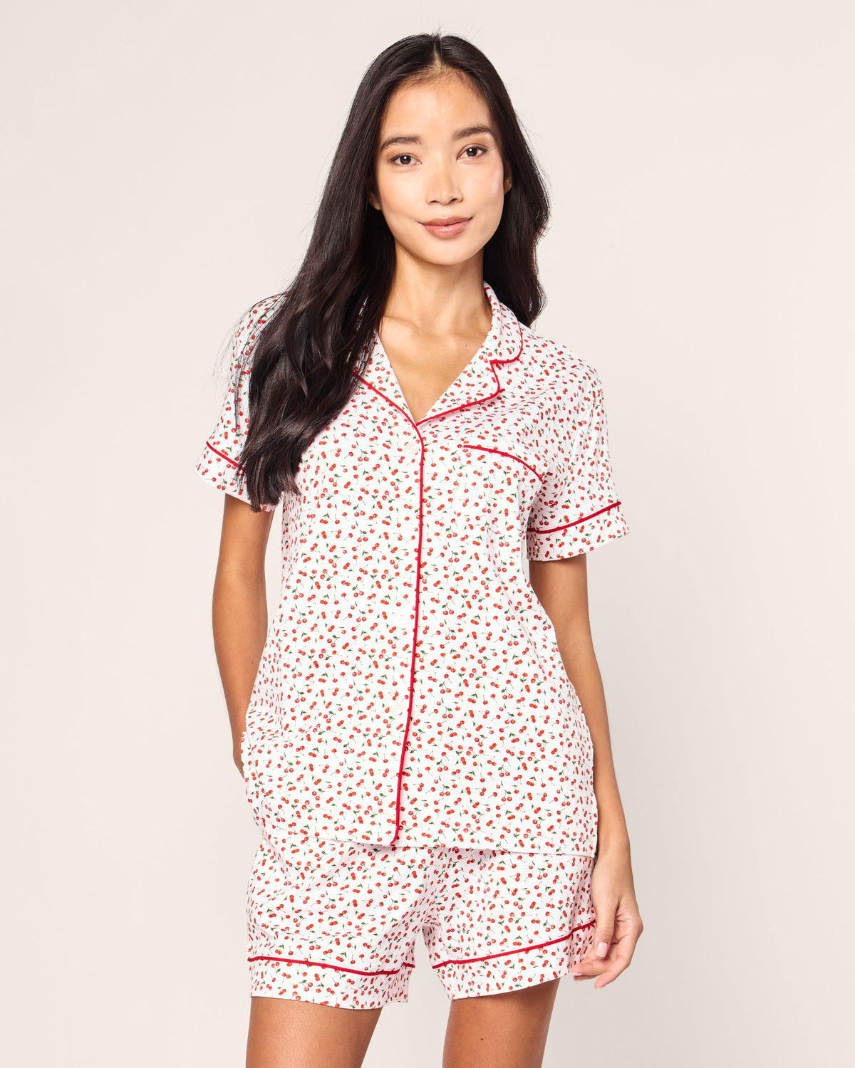 A person with long dark hair wears Petite Plumes Womens Pima Short Sleeve Short Set in Cherry Orchard, a white jersey knit pajama ensemble with a collared top, short sleeves, matching shorts, and red piping on a neutral background.