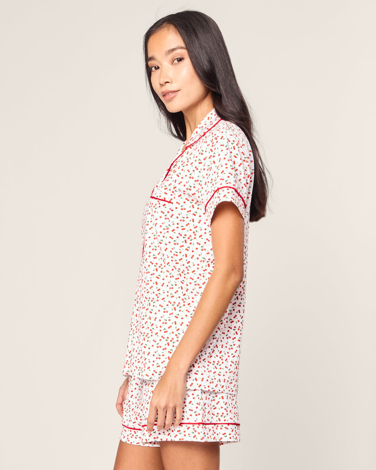 A person with long dark hair stands sideways in a Womens Pima Short Sleeve Short Set in Cherry Orchard by Petite Plume. The set features red floral patterns and red trim on a white background, with a short-sleeve top and matching shorts. The backdrop is plain and light-colored.