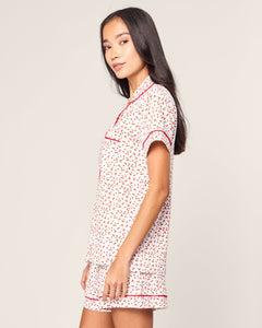 A person with long dark hair stands sideways in a Womens Pima Short Sleeve Short Set in Cherry Orchard by Petite Plume. The set features red floral patterns and red trim on a white background, with a short-sleeve top and matching shorts. The backdrop is plain and light-colored.
