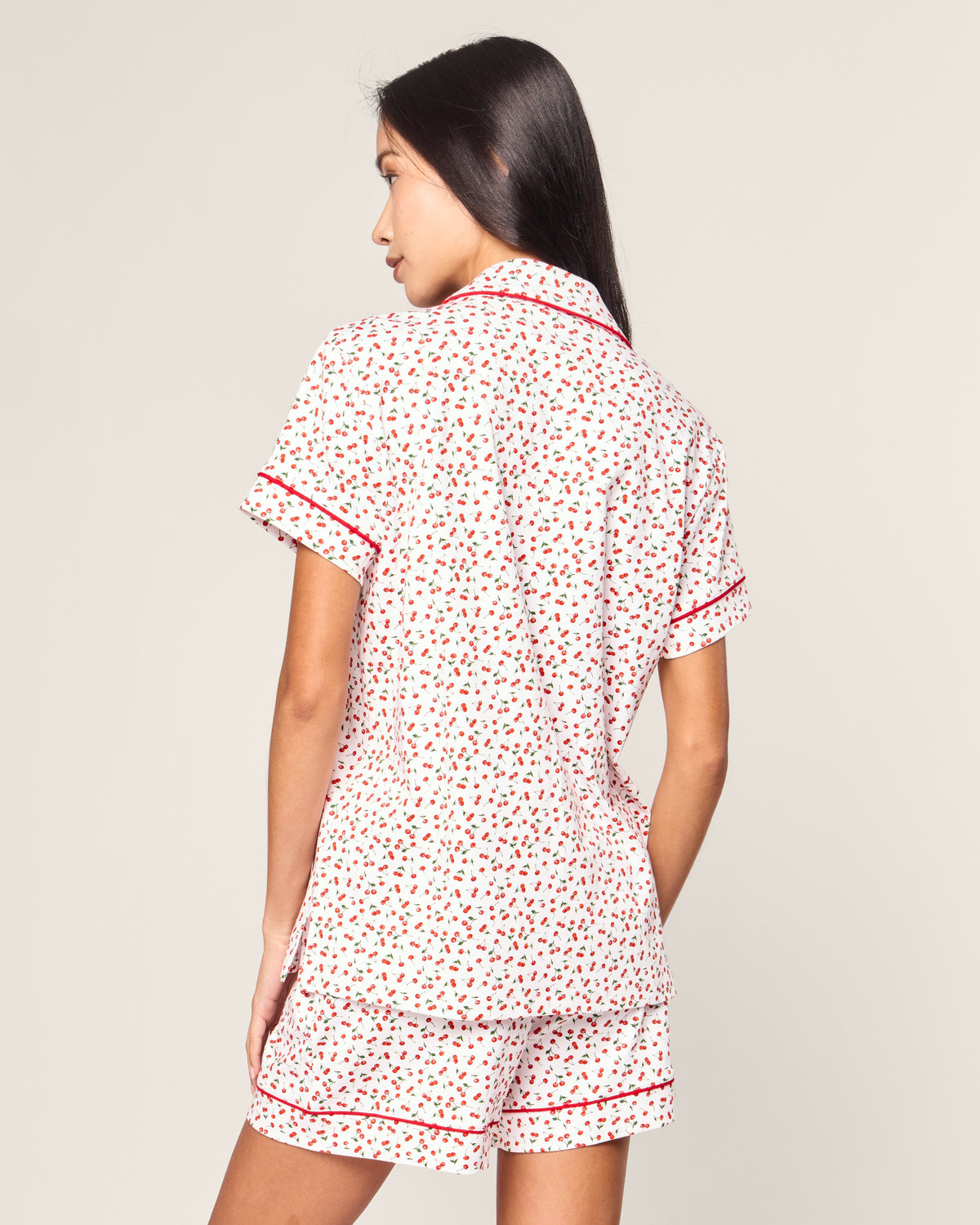 A woman with long dark hair faces away, showcasing the Petite Plume Womens Pima Short Sleeve Short Set in Cherry Orchard. The white pajama set features red heart patterns, a collar, and red trim.