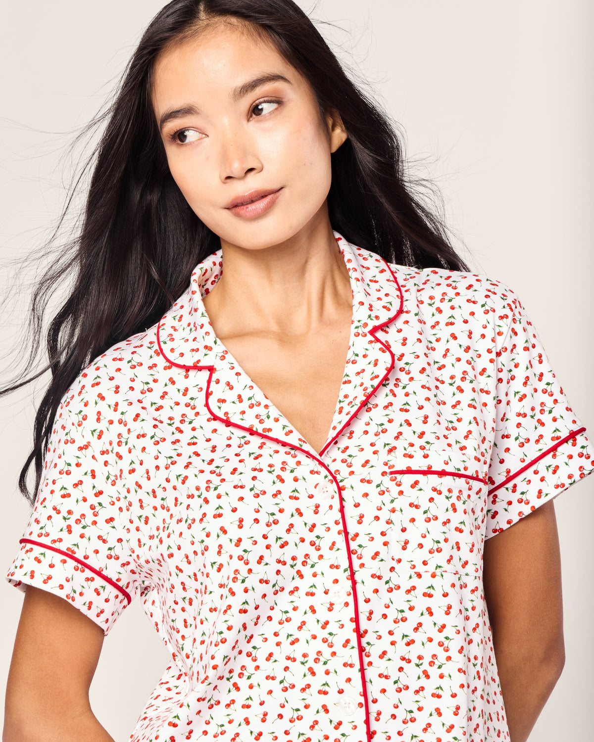 A woman with long, dark hair wears the Petite Plume Womens Pima Short Sleeve Short Set, displaying a Cherry Orchard print of red and black florals. The set is adorned with elegant red piping along the collar and sleeves as she gazes slightly to the side with a neutral expression.