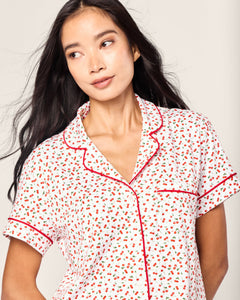 A woman with long, dark hair wears the Petite Plume Womens Pima Short Sleeve Short Set, displaying a Cherry Orchard print of red and black florals. The set is adorned with elegant red piping along the collar and sleeves as she gazes slightly to the side with a neutral expression.