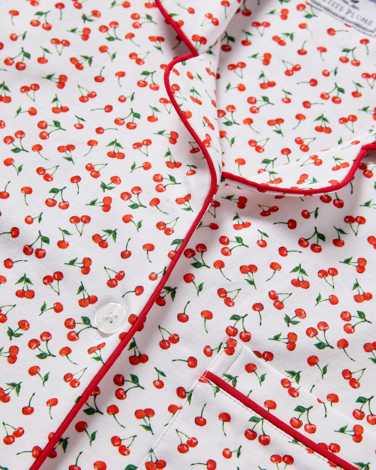 The Petite Plume Womens Pima Short Sleeve Short Set in Cherry Orchard showcases a print with small red cherries and green stems. It features red piping, a white button, and a pocket, all in soft jersey knit for comfort.