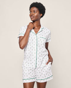 A person models the Womens Pima Short Sleeve Short Set in Palmier by Petite Plume. The white pajama set features a small pattern, green piping on the short-sleeved shirt and shorts, and embodies classic PJ comfort. One hand touches their neck while the other rests in a pocket.