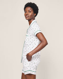 A person with short dark curly hair stands in a profile against a plain background, wearing the Petite Plume Womens Pima Short Sleeve Short Set in Palmier. The classic PJs are made of Peruvian Pima cotton and feature a small floral pattern with green piping, and they have one hand in their pocket.