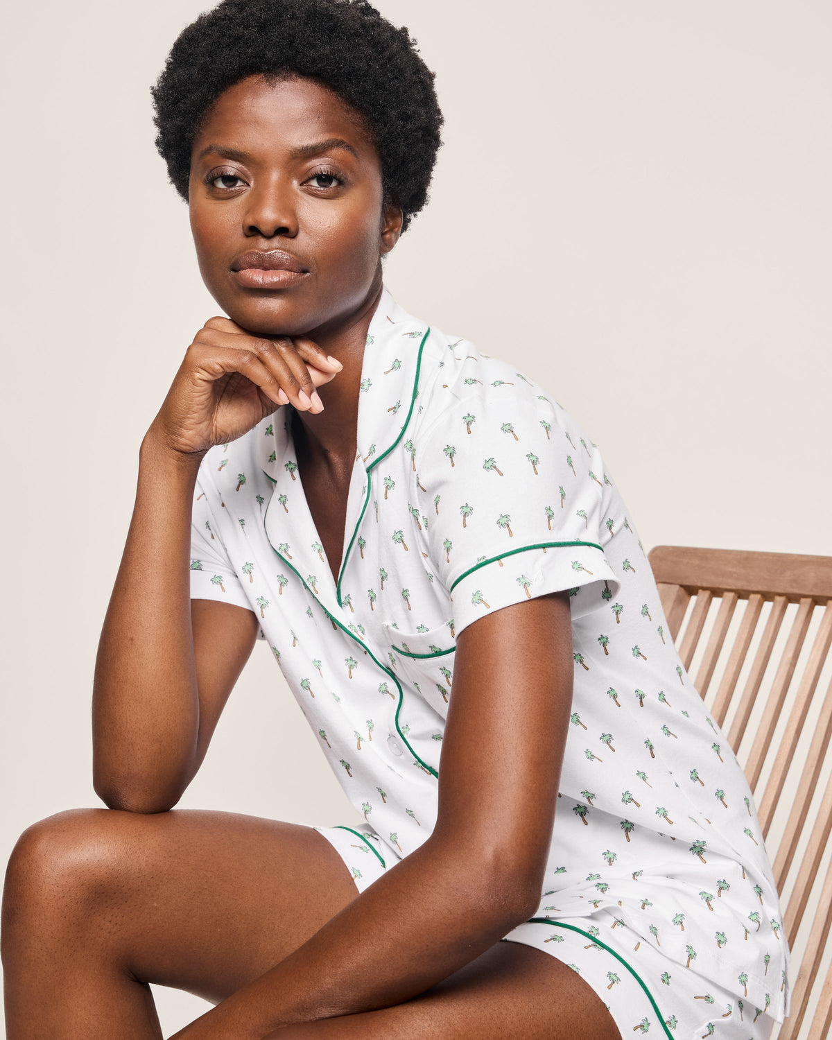 A person in a Womens Pima Short Sleeve Short Set in Palmier by Petite Plume, with short hair and a relaxed demeanor, sits on a wooden chair. They rest their chin on one hand, looking thoughtfully at the camera against a neutral background.