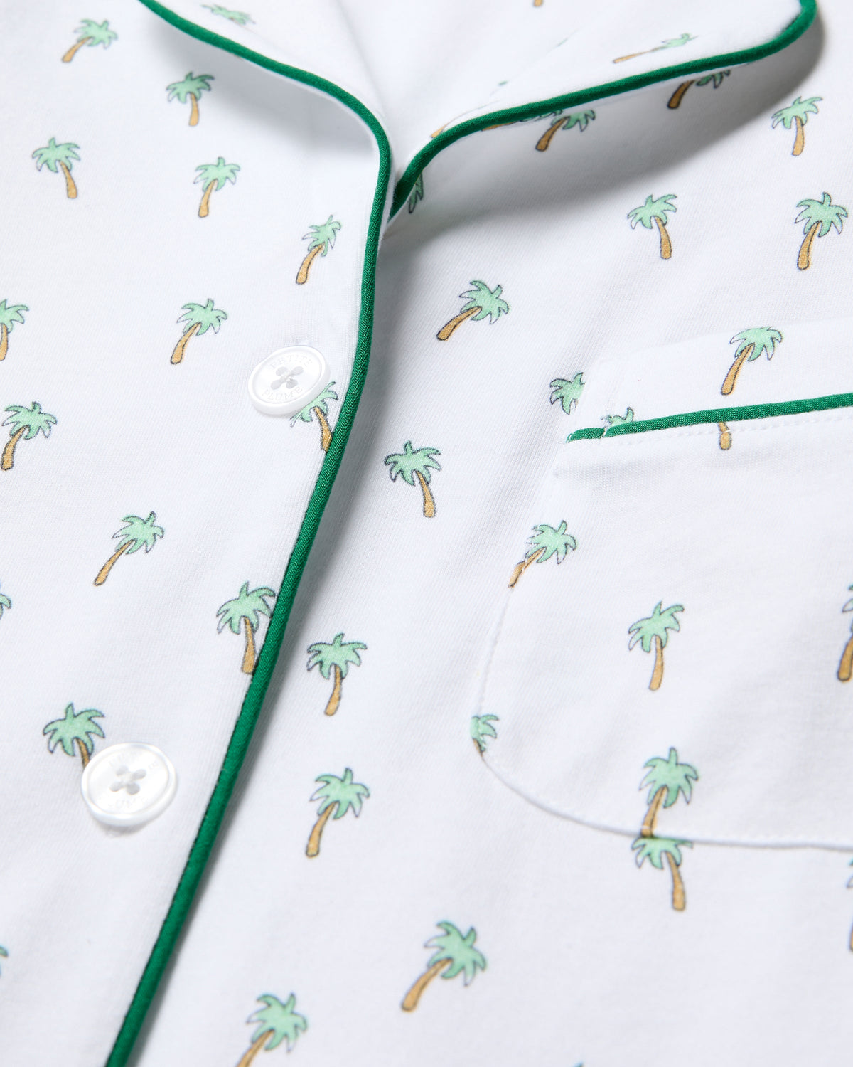 Close-up of the Womens Pima Short Sleeve Short Set in Palmier by Petite Plume, featuring a white shirt with palm tree prints and green piping. It has a right-side pocket and is fastened with elegant white buttons, delivering classic PJ comfort.