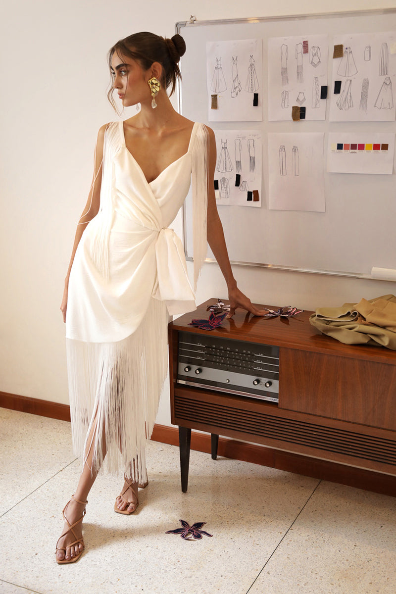 Three-quarter view of Parchita dress in white on model, fitted at the waist, showcasing draped V-neck, wrap tie and long fringe hem against a studio wall and vintage console