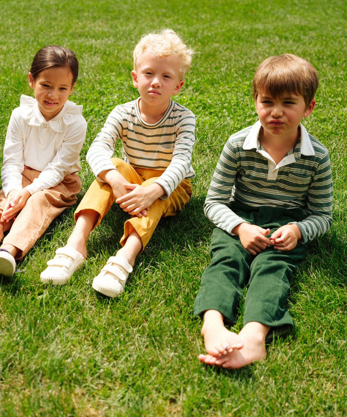 Boy wearing Parker Polo in Pine Twin Stripe, relaxed fit, sitting on grass with two other children in a sunny outdoor setting