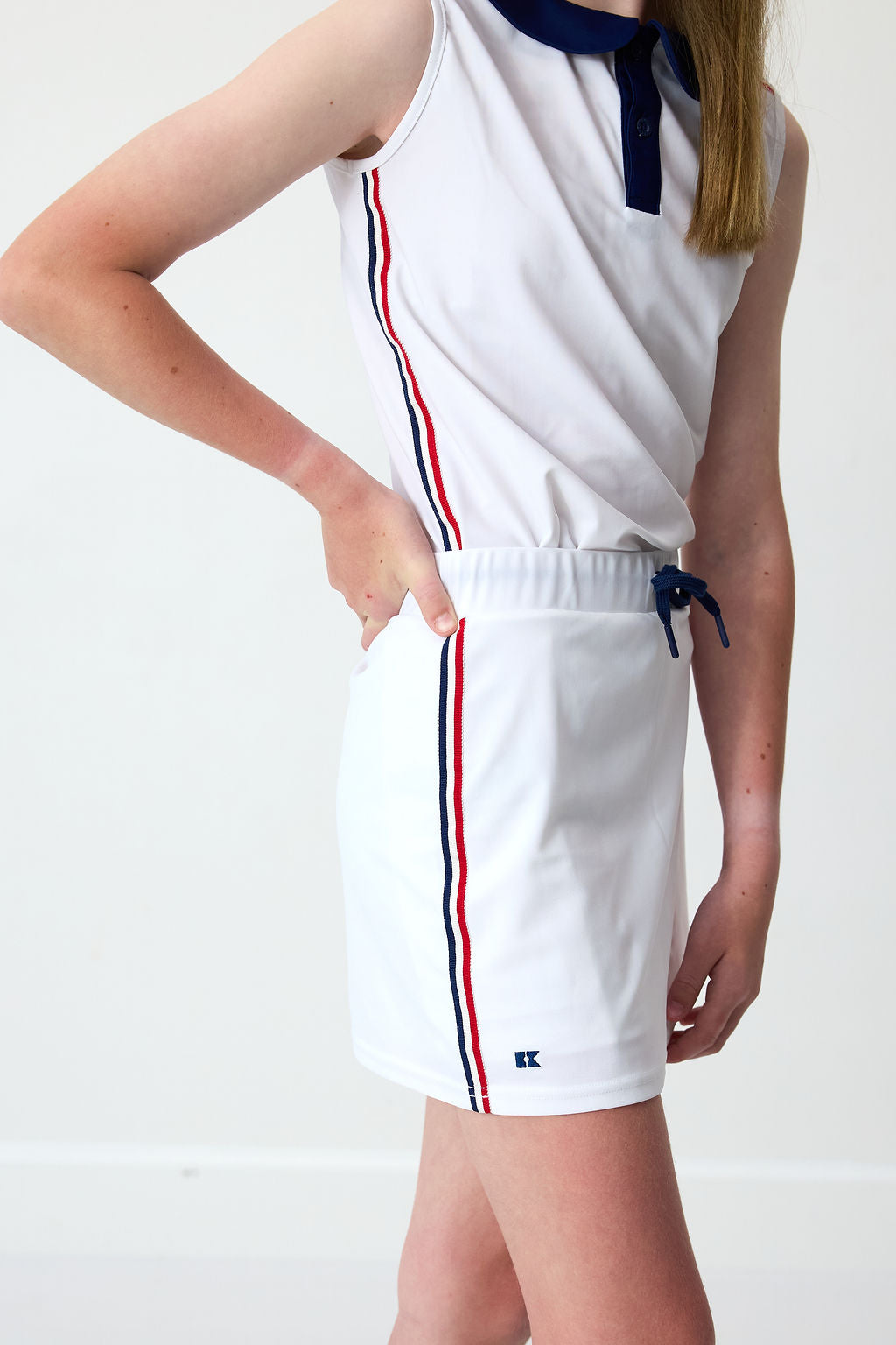 Side view of Liberty Love skort in white showing elastic waistband with navy drawstring, red-and-navy side stripes, and small logo on light background