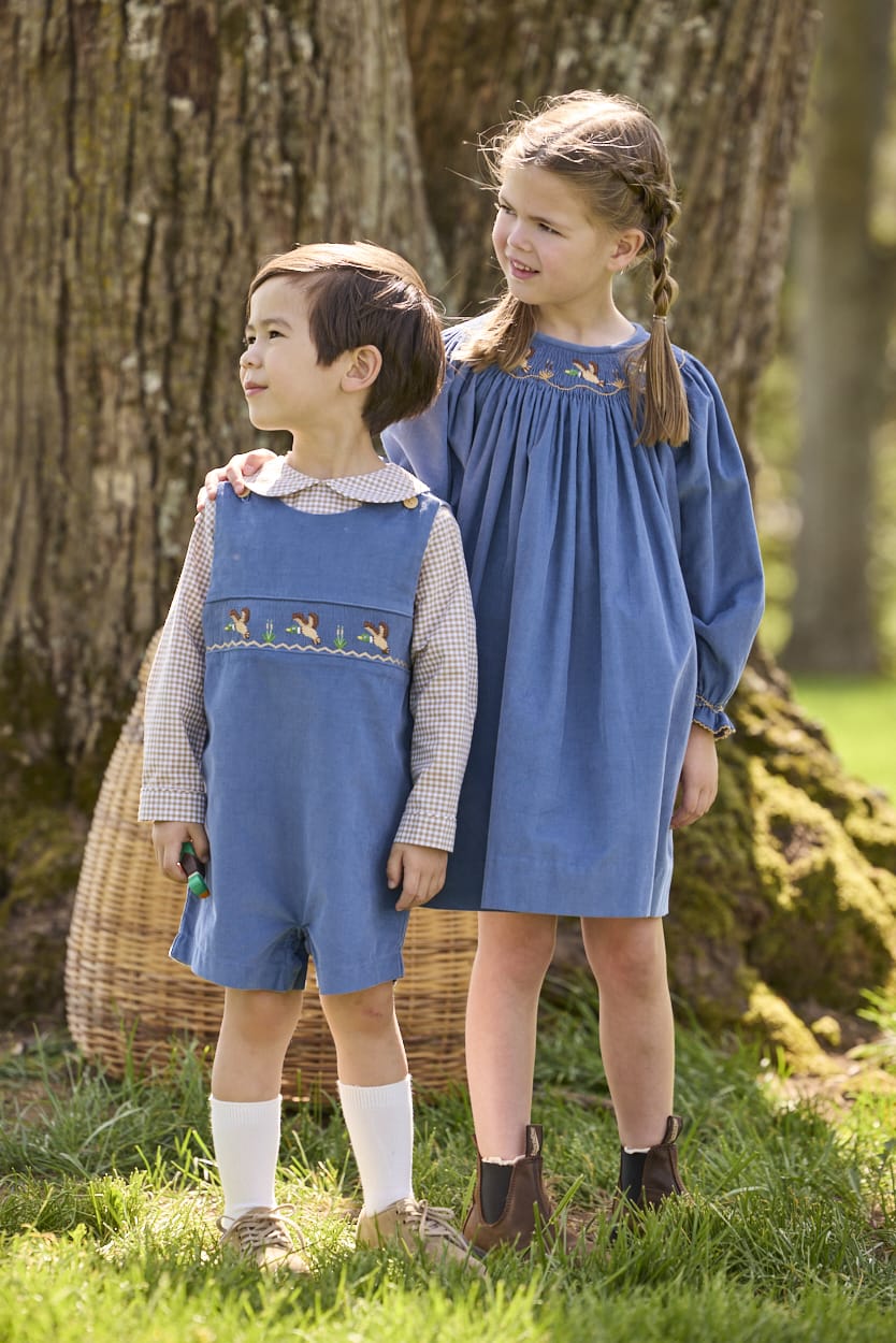 Little English traditional children's clothing.  Khaki Gingham peter pan shirt for toddler boys paired with stormy blue corduroy shortall with flying mallard smocking across chest. Long sleeve stormy blue corduroy dress with flying mallard collar around the neckline for little girls.