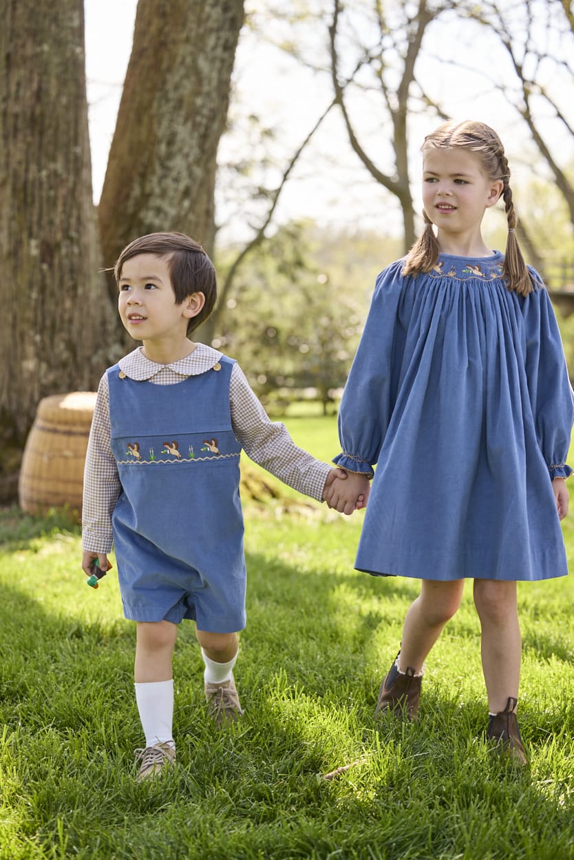 Little English traditional children's clothing.  Khaki Gingham peter pan shirt for toddler boys paired with stormy blue corduroy shortall with flying mallard smocking across chest. Long sleeve stormy blue corduroy dress with flying mallard collar around the neckline for little girls.
