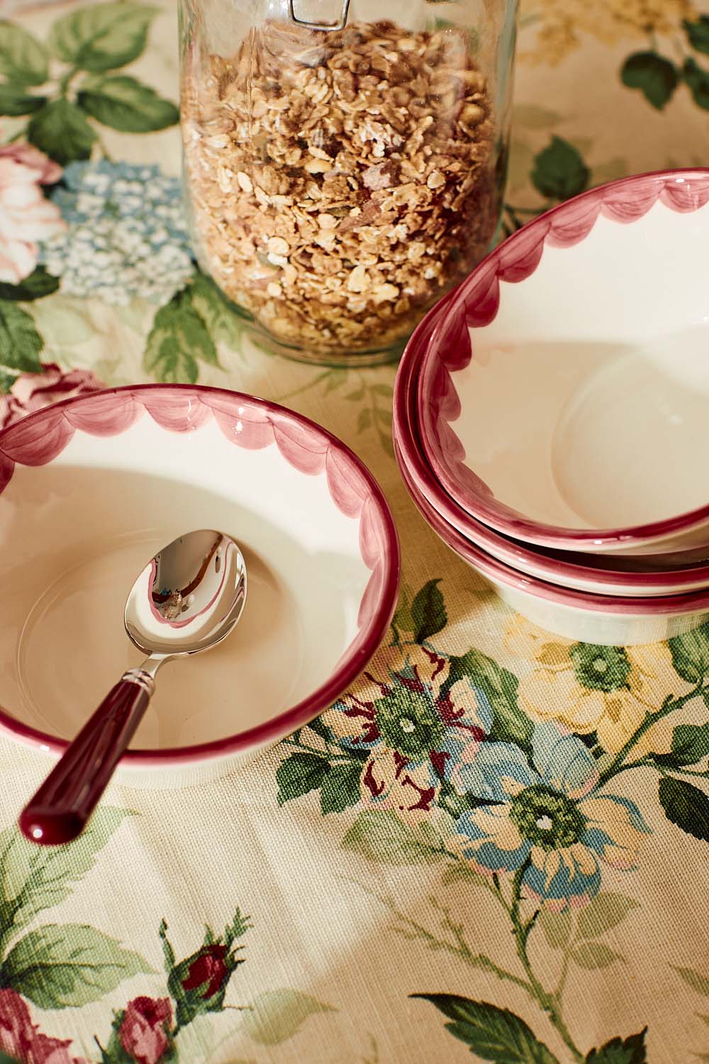 Top-down view of Pink Scallop Bowl showcasing scalloped hand-painted pink rim, stacked set with spoon and granola jar on floral tablecloth