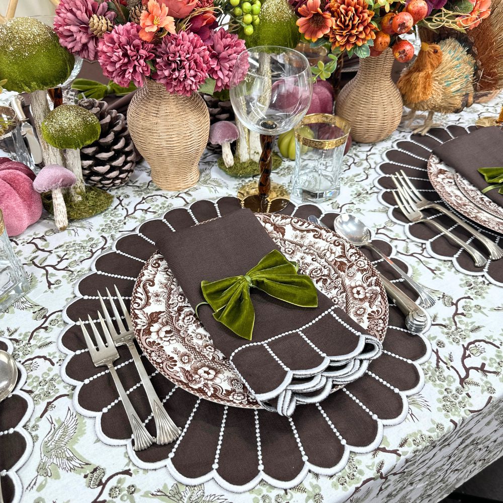 Decorative table setting with floral arrangements, plates, and cutlery on a toile sage patterned tablecloth with luxury linen placemats
