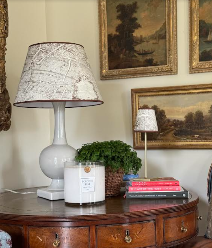 Two map-print Plan de Paris lamps on wooden side table with candle, potted fern, books, and framed paintings