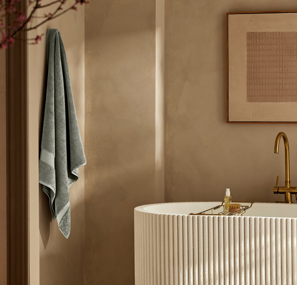 Plush Juniper bath towel hanging on a wall hook beside a ribbed freestanding bathtub in a warm beige bathroom
