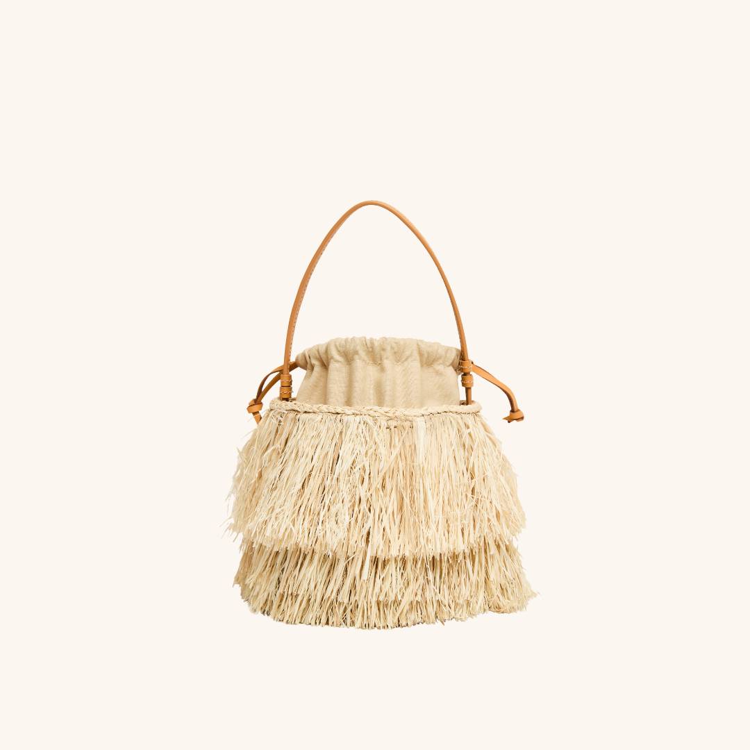 Front view of The Harbour Island Bucket in Natural showcasing layered straw fringe and tan leather handle on a light beige background