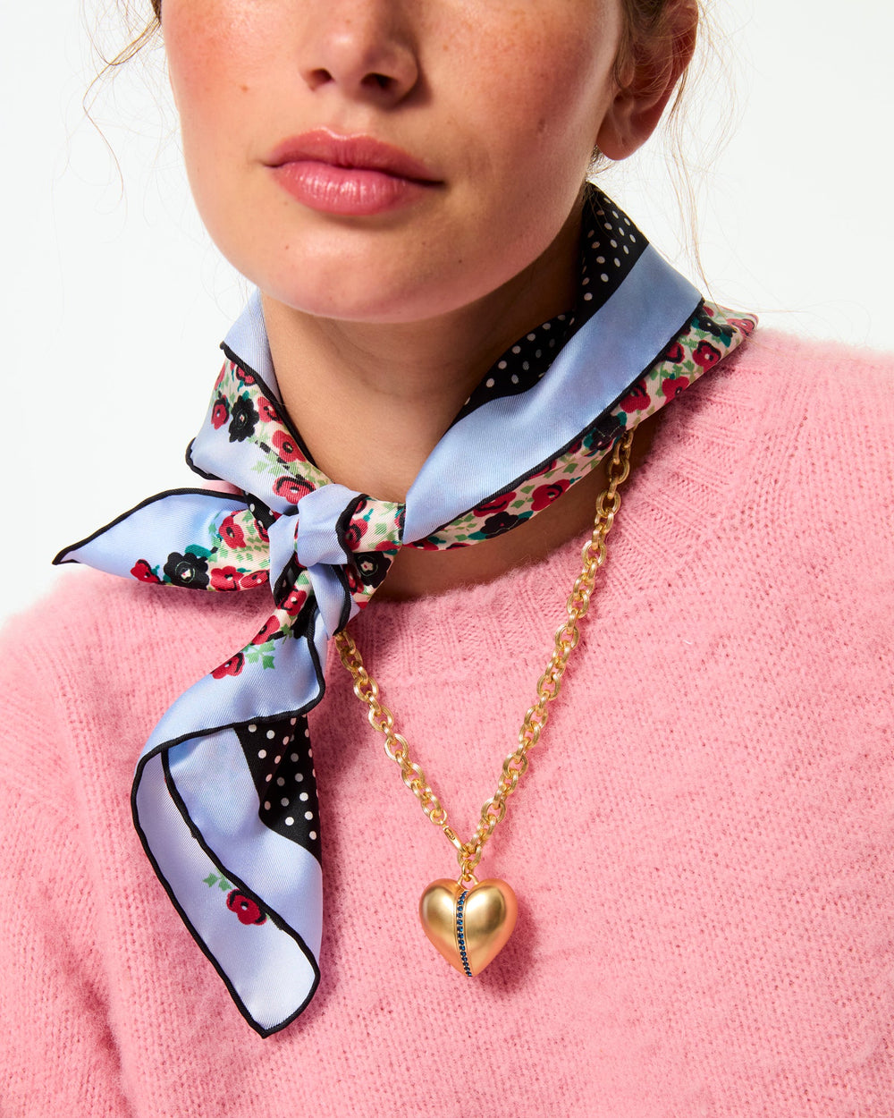 Close-up of Sapphire Stripe Puffy Heart pendant, satin gold-tone heart with sapphire stripe on chain worn over pink sweater