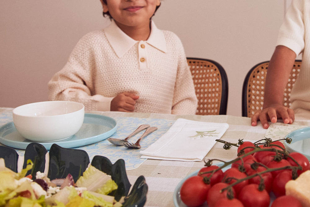 Two children seated at a dining table with an ivory ceramic bowl on a blue plate, salad and vine tomatoes in the foreground