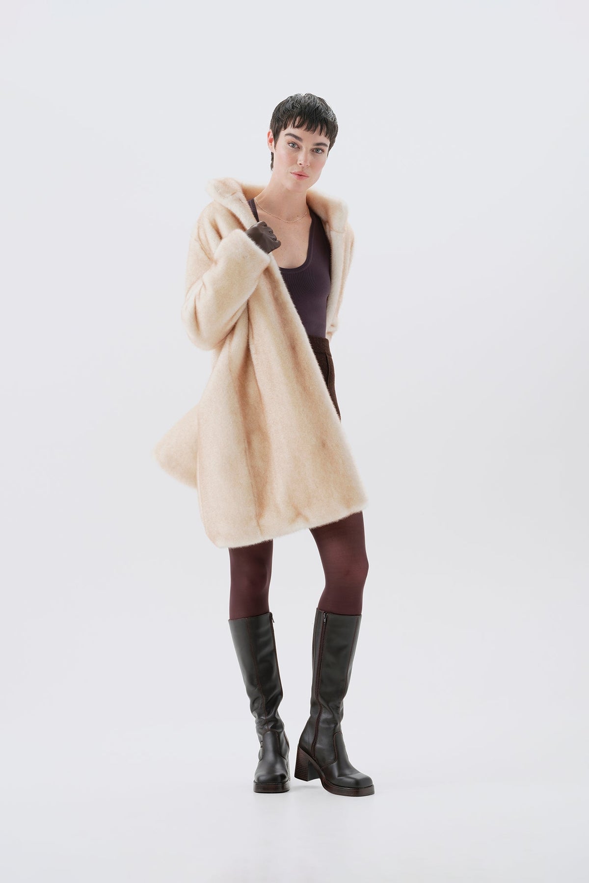 Model wearing a beige faux fur coat, dark pants, and black boots on a white background