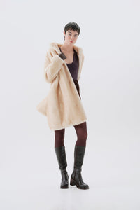Model wearing a beige faux fur coat, dark pants, and black boots on a white background