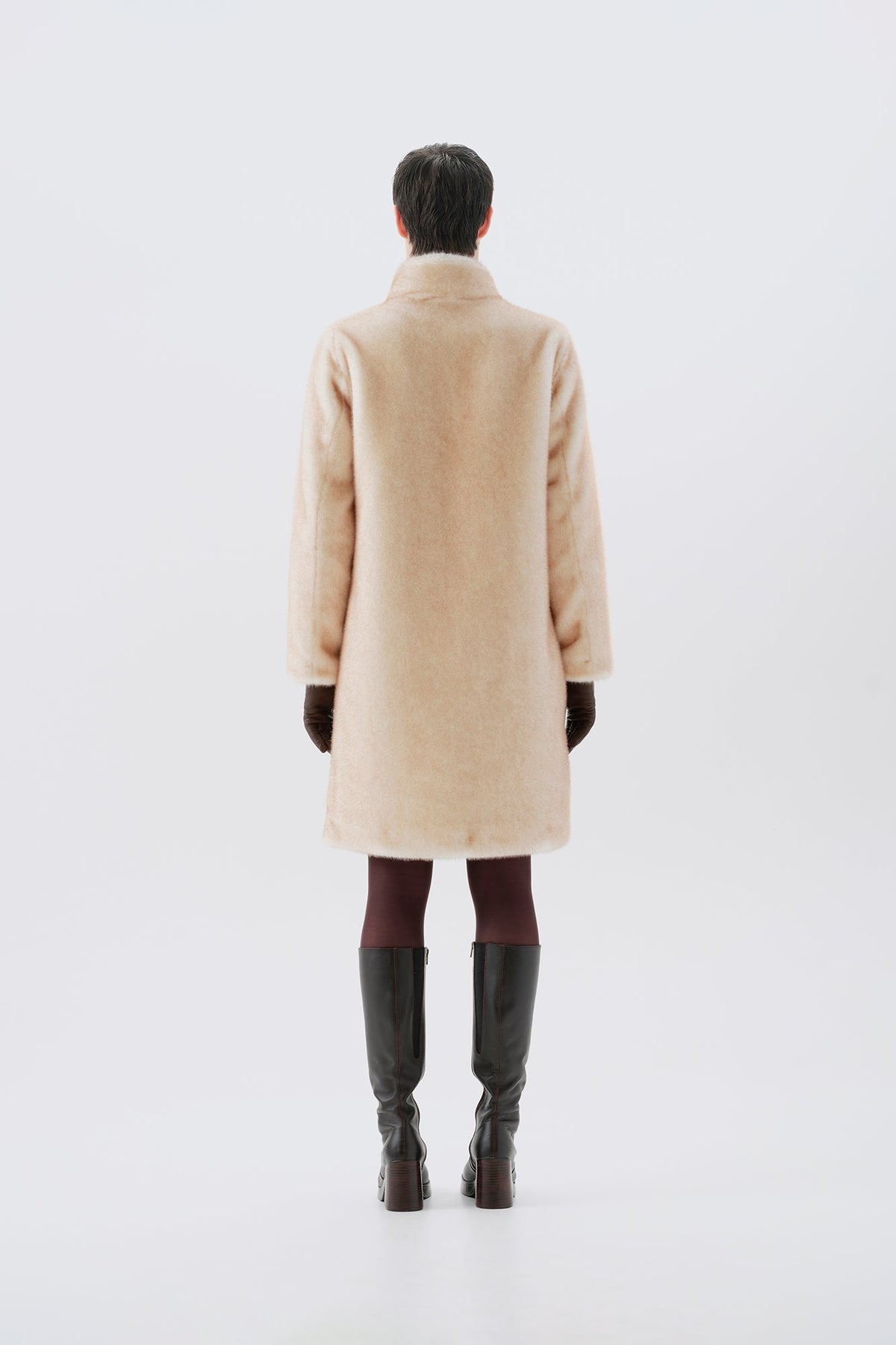 Model pictured from the back wearing a beige faux fur coat, dark pants, and black boots on a white background
