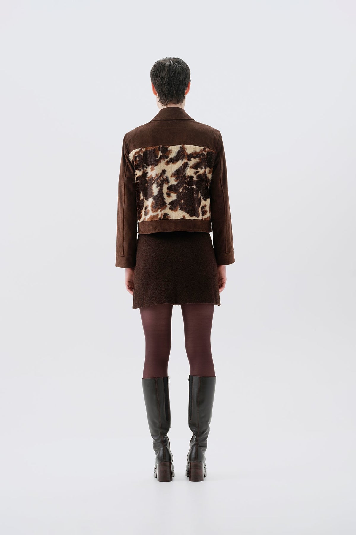 Model pictured from the back wearing a faux fur cow print jacket with brown vegan suede sleeves on a white background
