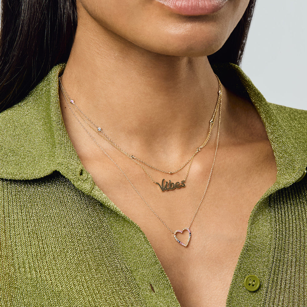Close-up of Rainbow Sapphire Heart Necklace on model showing a multicolor sapphire heart pendant on a 14ct gold chain layered over a green knit top