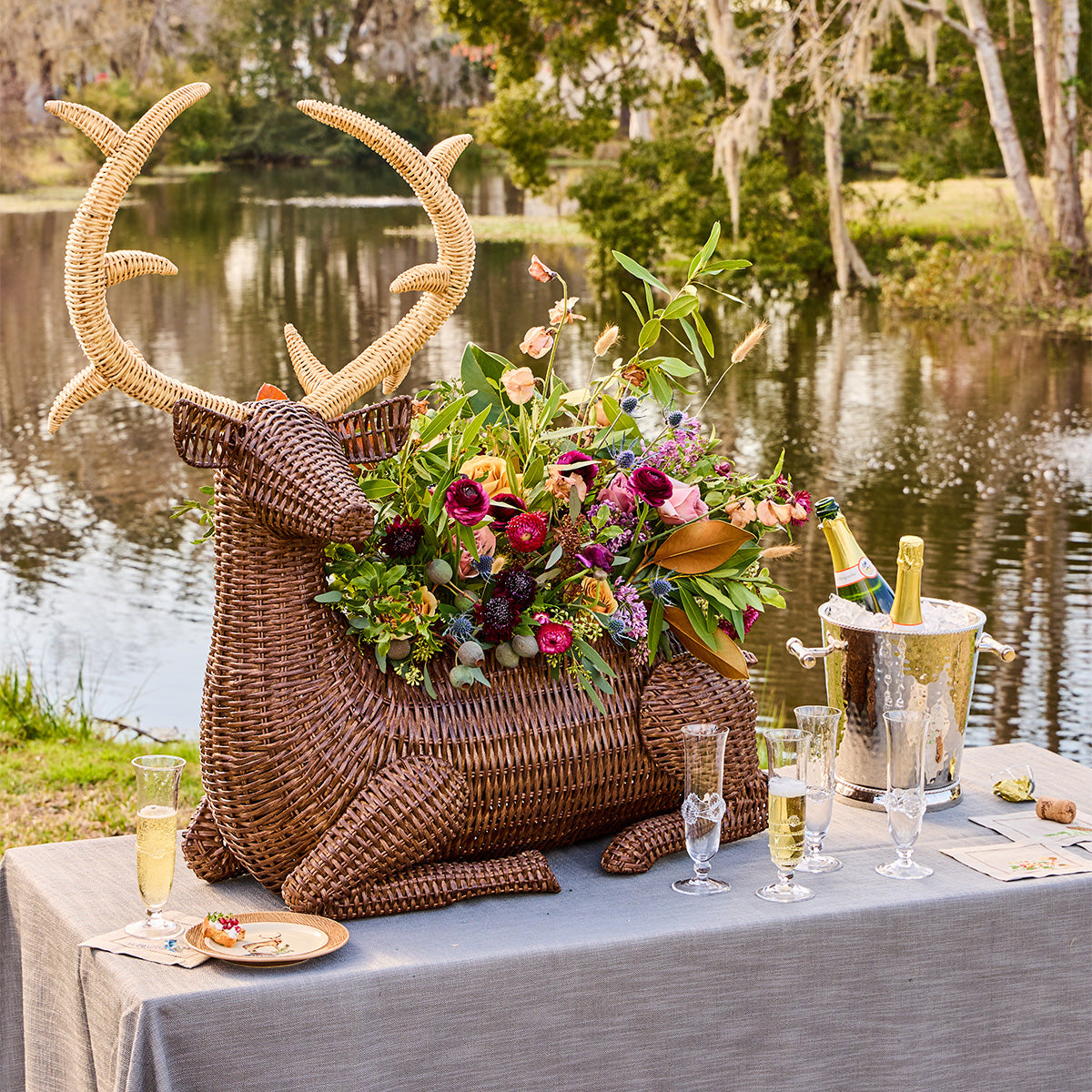 Front view of Devon Willow Stag Party Bucket showcasing woven rattan stag and floral-filled basin on a lakeside table