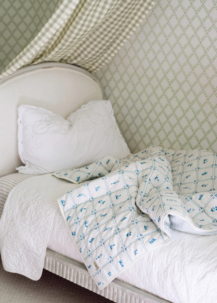 Quilt draped on bed showing Delft Quilt’s blue grid motif and rice stitching with white headboard and green wallpaper