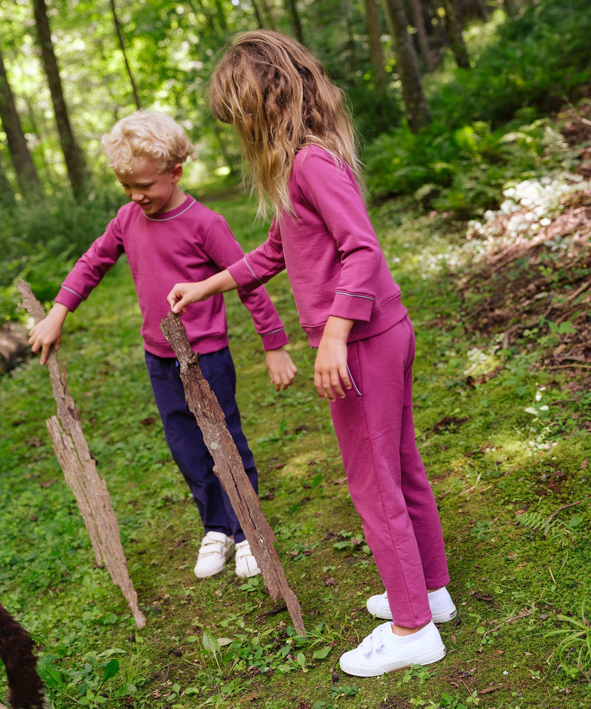 Two children wearing blackberry Remy sweatshirts playing with sticks on a grassy forest path