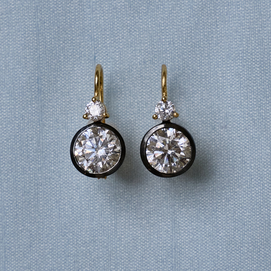 Front view of Rhodium Double Bezel Drop Earrings showcasing round faceted stones with a light blue textile background